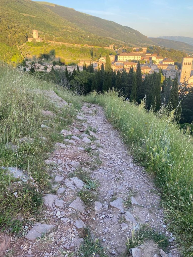 Sentiero sterrato tra l’erba con un paese illuminato dal sole al tramonto sullo sfondo