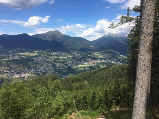 Vista panoramica sulla Val di Fiemme dall’alto, con boschi di abeti, paesi sul fondovalle e montagne sullo sfondo