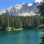 Lago di Carezza in una giornata soleggiata, con bosco di abeti e le cime dei Campanili del Latemar sullo sfondo
