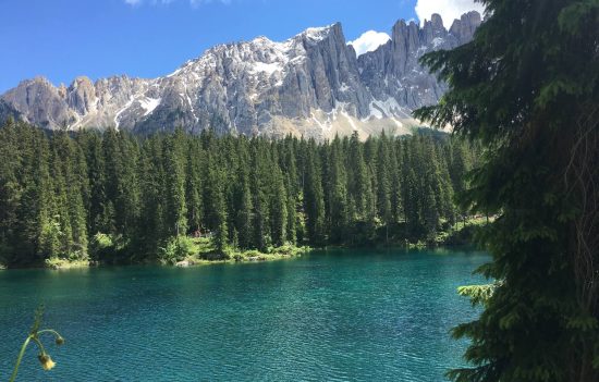 Lago di Carezza in una giornata soleggiata, con bosco di abeti e le cime dei Campanili del Latemar sullo sfondo