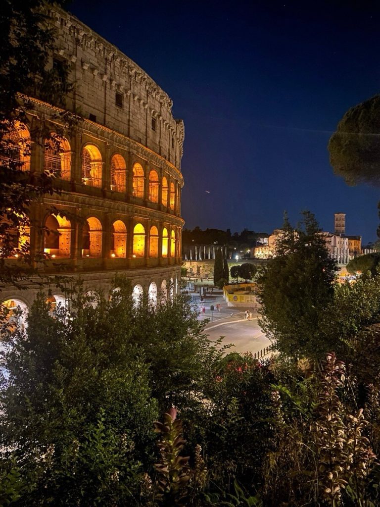 Il Colosseo illuminato di sera, visto di lato tra alberi e cespugli, con il Foro Romano sullo sfondo.